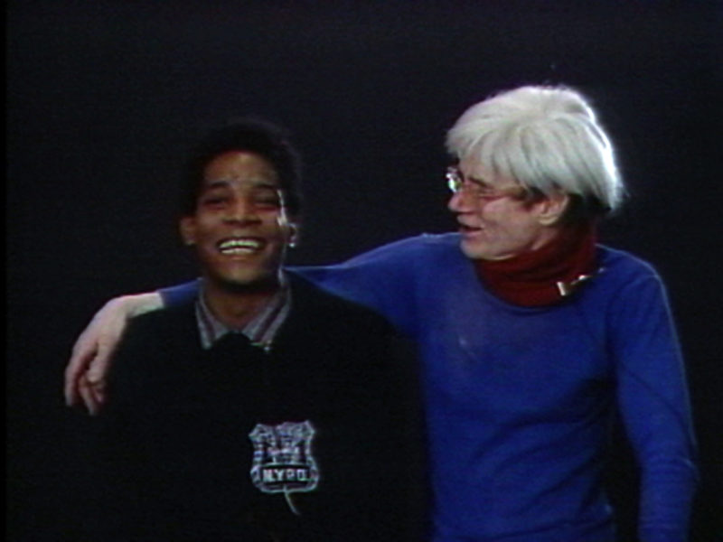 Jean-Michel Basquiat faces forward and smiles as Andy Warhol drapes his arm over his shoulders and looks at him