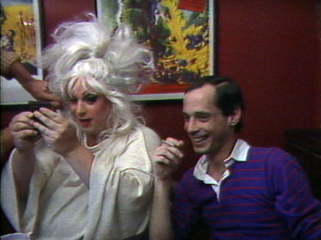 Video still of Divine looking at herself in a pocket mirror, next to John Water who is smiling and holding a cigarette