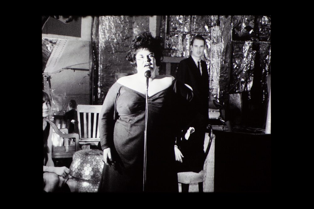 A black and white film still of Tally Brown singing behind a mic is projected onto a wall