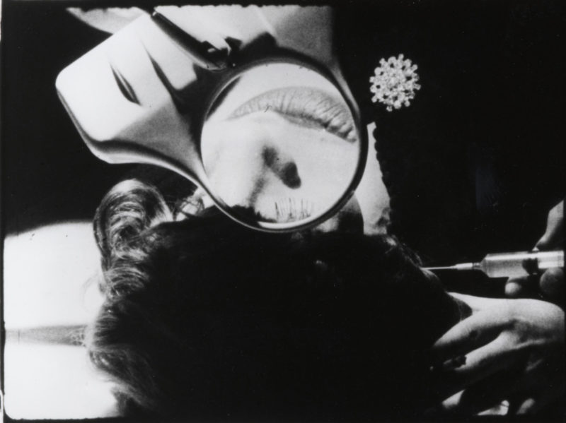A somewhat abstract black and white film still, in which a nose and lips can be seen through a magnifying lens, and a syringe is being held in two hands