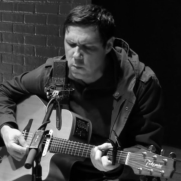 Man with guitar strums while singing into a microphone