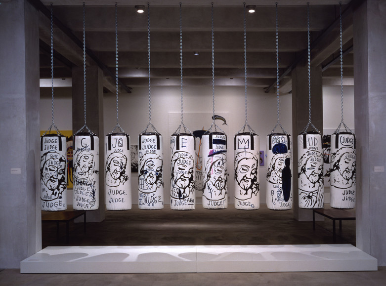 Ten white punching bags hang from chains in a row in the middle of a gallery. The punching bags have Jesus' face and the word JUDGE painted on them in black in varying sizes and positions.