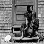 Woman with guitar sits on the steps of a cottage with weathered split-shake shingles.
