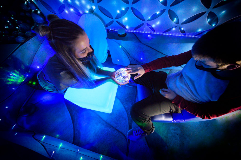 In an enclosed, dim space, one person is sitting in a chair. The room is lit in vibrant blue and green with a lot of lights simulating starlight on the walls. A second person is popping out of a set piece on the far end of the space and extending a glowing orb to the sitting person, who is touching it gingerly with a couple of fingers. Each person is wearing a mask covering their nose and mouth. This is shown from an overhead angle.