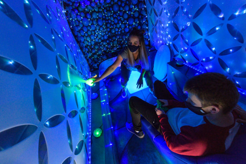 In an enclosed, dim space, one person is sitting in a chair and a second person is sitting on a glowing cube. The room is lit in vibrant blue and green with a lot of lights simulating starlight on the walls. The two people are seated across the room from each other. The picture is taken from behind one person, focusing on the other’s movements. She is shining a green light on the wall. Each person is wearing a mask covering their nose and mouth.