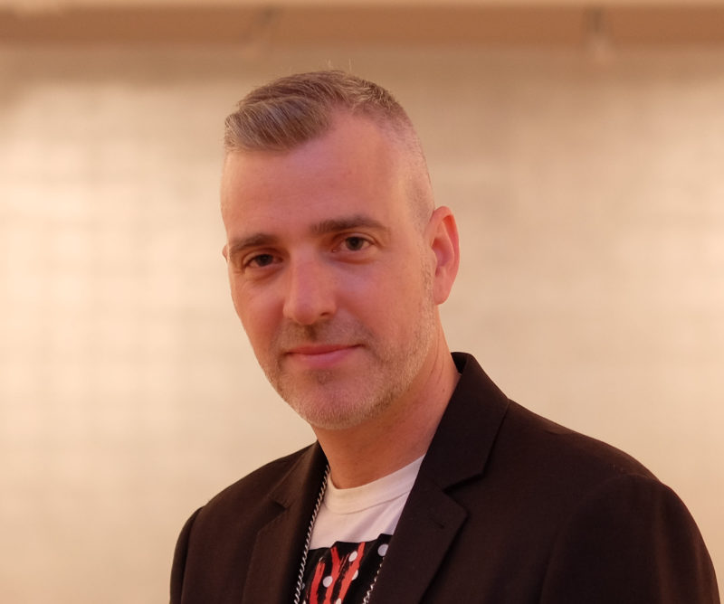 A picture of a person in a t-shirt and black jacket in front of a white, tile background looking at the camera.