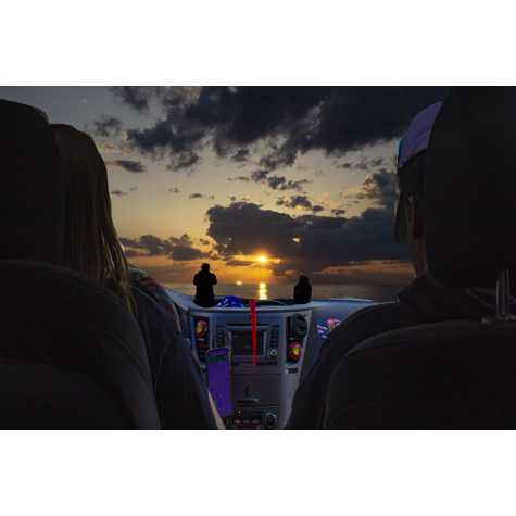 Two people sitting in car, we see the back of their heads against a car seat headrest as they look out into a sunset over the beach