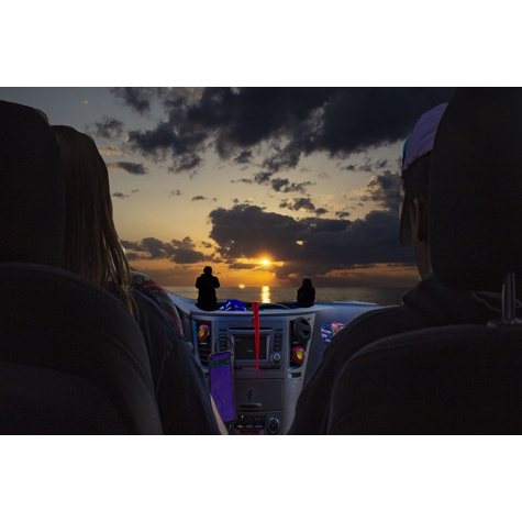 Two people sitting in car, we see the back of their heads against a car seat headrest as they look out into a sunset over the beach