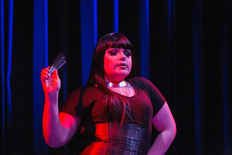 A young drag queen wearing all black with a long black wig and exaggerated makeup, pictured from the waist up. She is holding a folded fan in her right hand as she sings. A pink colored light shines on her while standing in front of dark blue stage curtains.