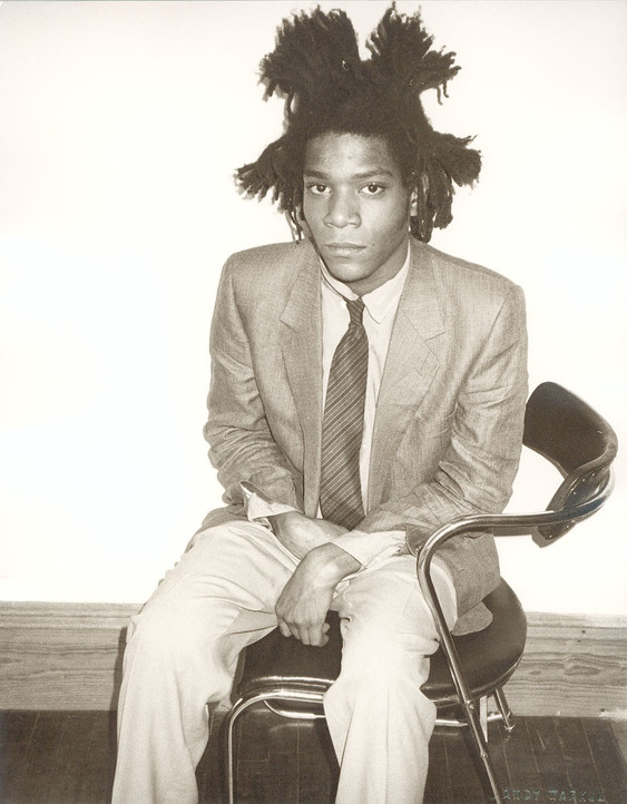 Black and white photograph of Jean-Michel Basquiat wearing a suit and sitting in a chair.