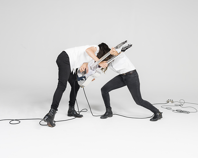 Two similar looking people, wearing white shirts and black pants, are struggling to play a silver double sided guitar. Their heads touch and it appears to mirror one another.