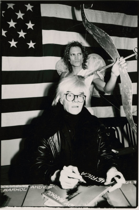 Warhol, seated at a table signing copies of his book 'America,' against the backdrop of an American flag and a man dressed as an angel.