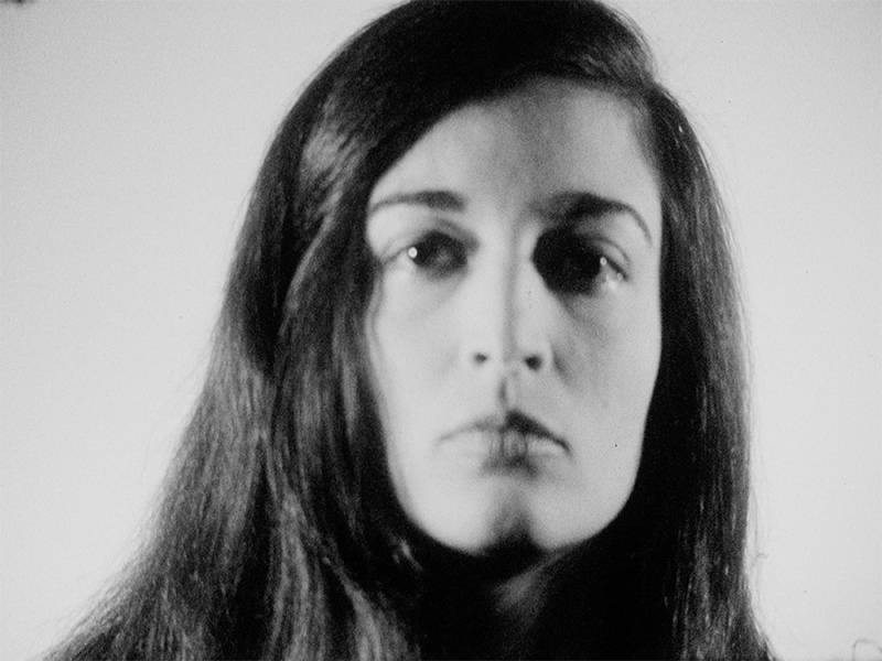 A black and white film still of a closeup view on the face of a person with long black hair who looks straight ahead with their eyes open.
