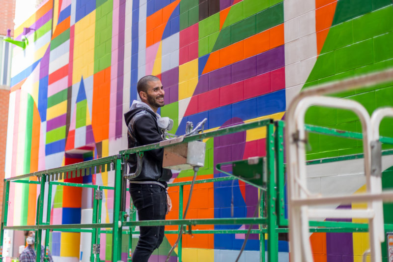 A smiling person, wearing black jeans and a black jacket, standing in front of a colorful wall with geometric patterns.