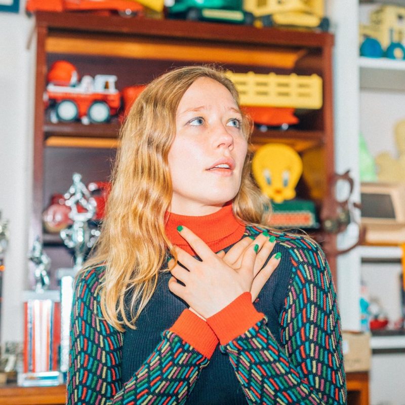 A person with long, blond hair stands in front of a shelf with various toys on it. She has her arms crossed on her chest and is looking off to the right side of the frame.