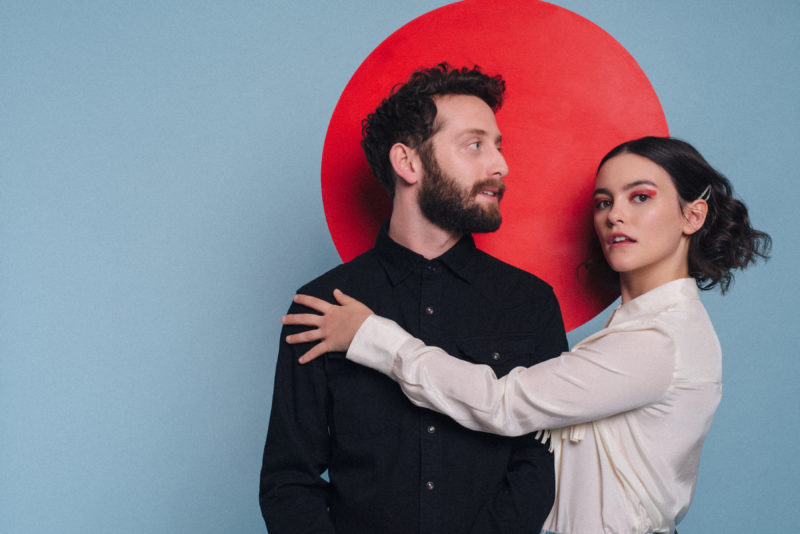 A person in a long sleeve white shirt is holding a person in a long sleeve black shirt in front of a red circle painted on a gray wall.