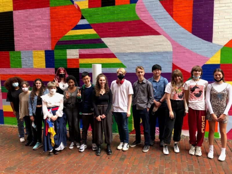 A group of 14 teenagers stand in a line in front of a colorful wall.