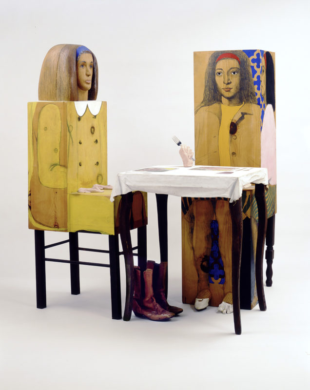 Two humanoid sculptures sitting at a table. Sculptures are a combination of sculpted features and illustrations on flat surfaces.