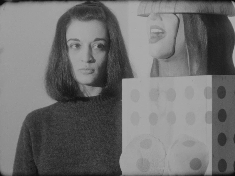 A black and white film still of Marisol looking at a sculpture. The sculpture is of the lower half of a person's head, sandwiched between abstract forms.