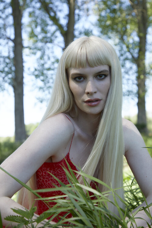 A person with long, blonde hair and wearing a red dress is looking towards the camera. There is a green plant in the foreground and trees in the background.
