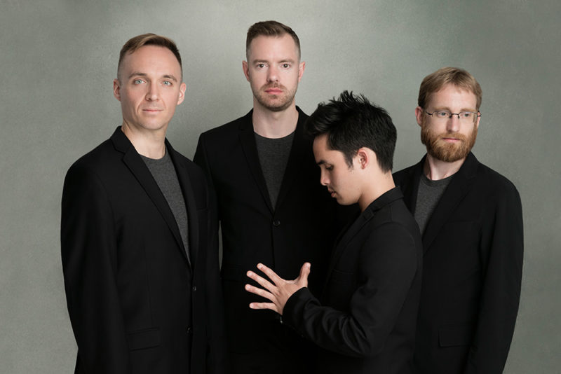 Four people wearing black suit coats over a gray shirt are standing in front of a gray background. One of them is standing sideways and has their hand with fingers stretched out in front of them.