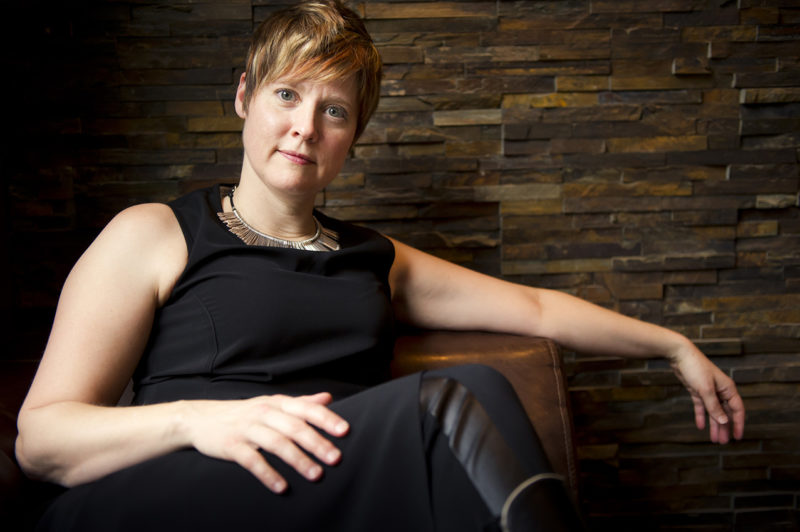 A person wearing a black dress sits on a tan chair in front of a stone wall.