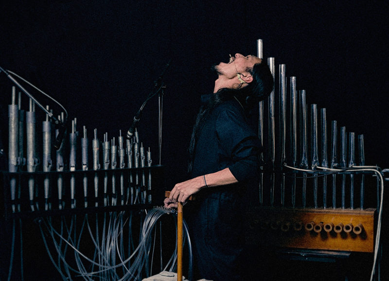 A person wearing all black stands in-between instruments made with many pipes. They are looking upward with their mouth wide open.
