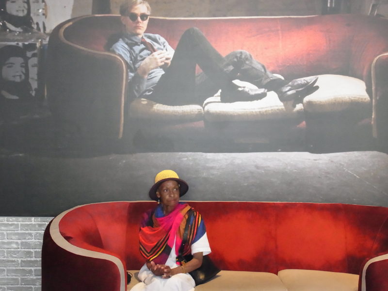 A women in a colorful dress sitting on a couch underneath an image of Andy Warhol