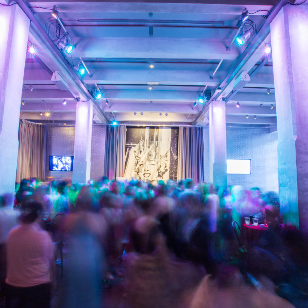 A group of people dancing and sitting at tables. On the wall behind the group, a picture of Andy Warhol holding up a large acetate of Marilyn Monroe’s face is visible.
