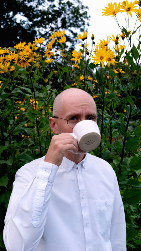 A person wearing a white, button down shirt stands in front of sunflowers while drinking out of a white mug.