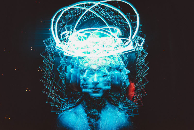 Multiple exposure photograph of a face turning side to side in a blue tint while in costume and makeup in front of a black background.