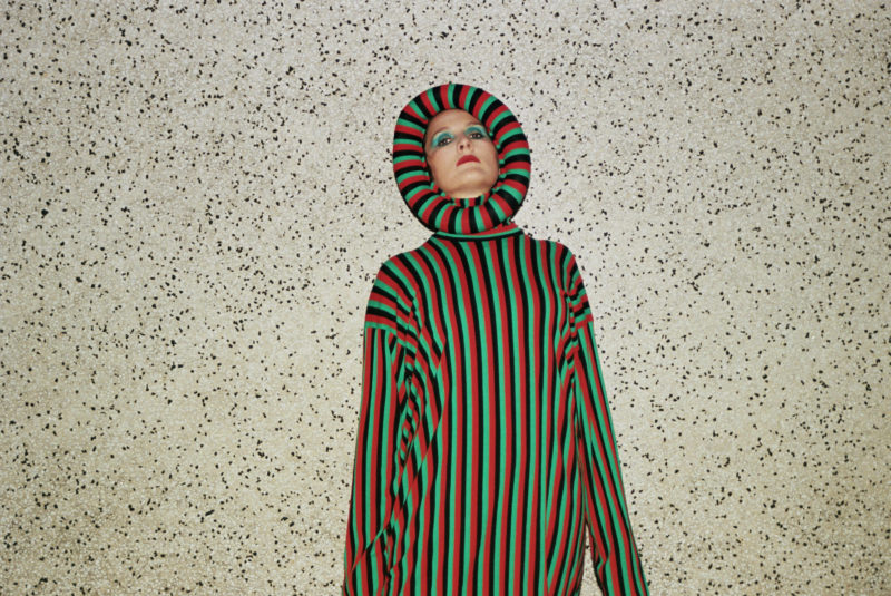 A person standing in front of a white wall speckled with black spots. They are standing straight up and wearing an outfit that covers everything except for their face. The outfit has straight, red, green, and black vertical lines.