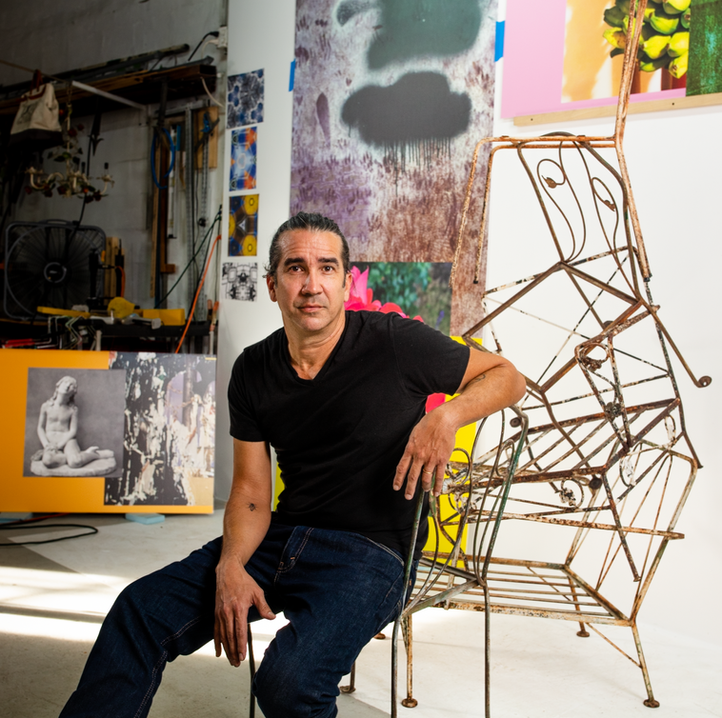 Man sits in chair next to a sculpture and with paintings hanging behind him on the walls of his studio.