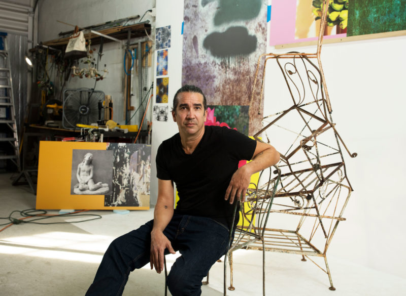Man sits in chair next to a sculpture and with paintings hanging behind him on the walls of his studio.