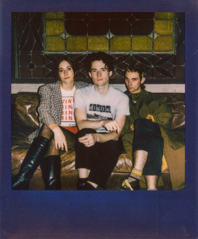 Three people sit on a couch in front of a stained glass window. They are all looking towards the camera.