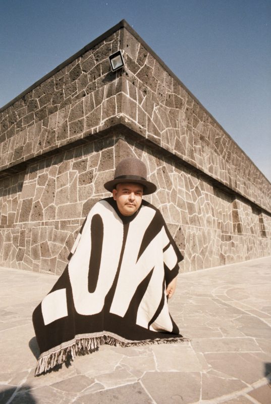 A person wearing a large, brown hat is outside in front of a stone building on a stone walkway leaning down as they are covered with a blanket so that only their face and left hand are showing.