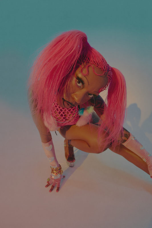A closeup of a person as they are bent down on the floor. They have pink hair in pig tails, are wearing pink fish net-style clothing, pink gloves and colorful bangle bracelets.