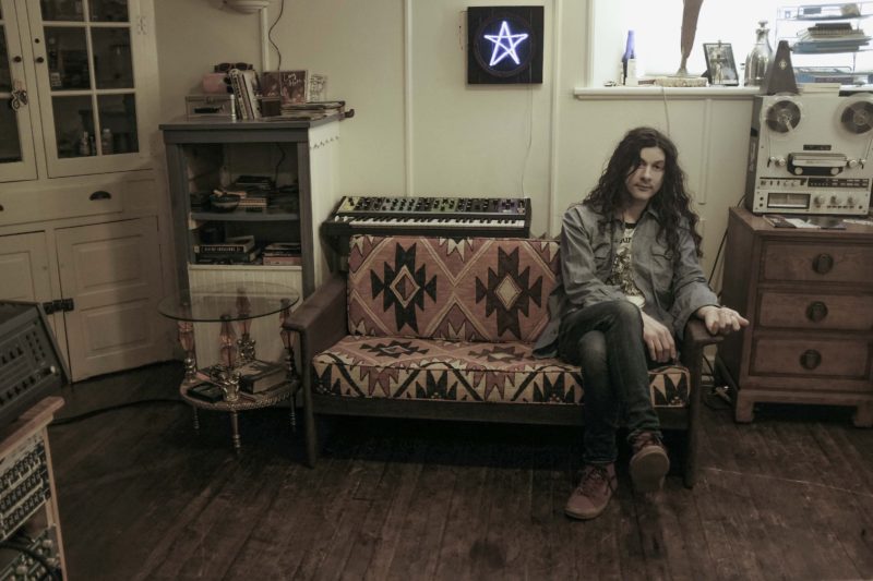 A person with long, brown hair sits on the end of a couch in a room with hardwood floors, a keyboard behind them, and studio equipment in various areas of the room.