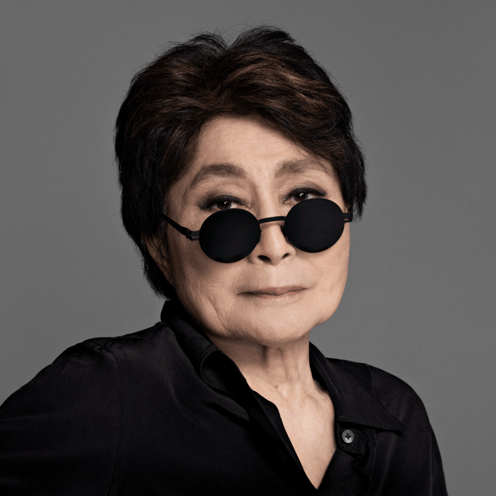 Woman with black shirt and black round sunglasses looks at the camera.