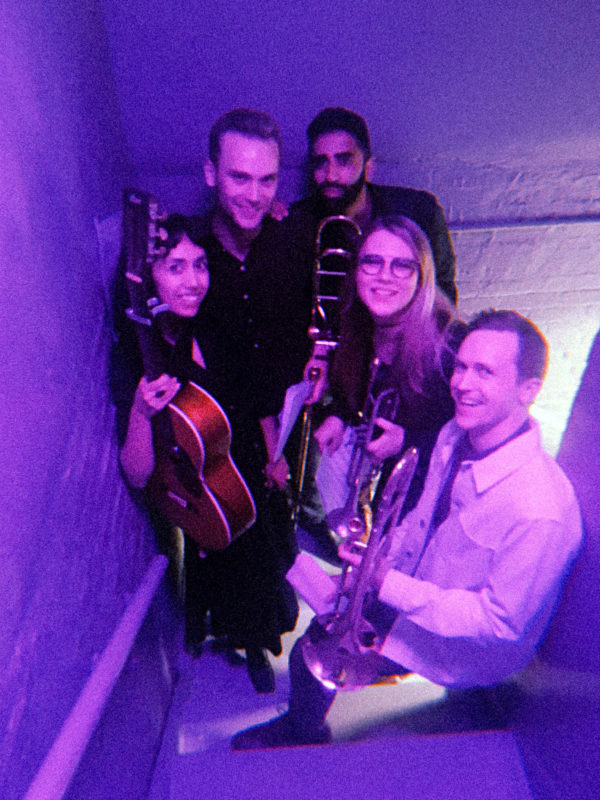 A group of five people holding various instruments stand on steps. There is a purple light shining over all of them.