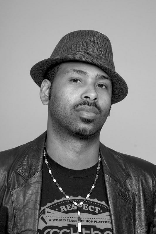 Black and white photograph of a person with a goatee and mustache looking towards the viewer, wearing a hat, leather-like jacket black t-shirt, and necklace.