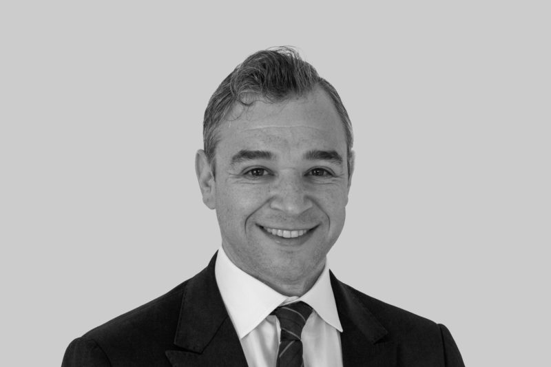 Black and white photograph of a person in a suit and tie looking towards the viewer and smiling in front of a light background.