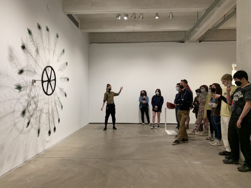 Group photo of teens on tour in a gallery at The Andy Warhol Museum, in front of Paola Pivi artwork of peacock feathers attached to a bicycle wheel shaped motor.