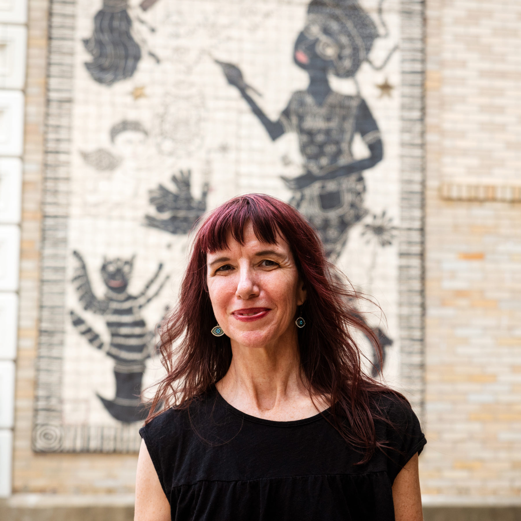 Artist stands in front of tile mural installed on a brick wall.