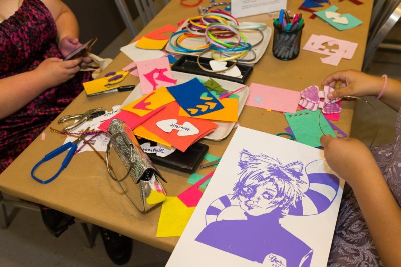 Two teens work on art projects in the Warhol’s studio space. There are colorful pieces of paper on the tables that the young people are using to create cat ears for headbands. A screen printed image of Andy Warhol as the Cheshire cat sits on the table in the foreground.