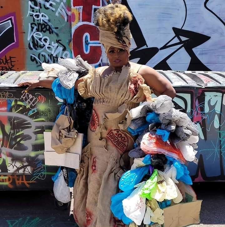 Drag Queen, Akasha L. Van-Cartier, stands posing in front of a wall and a dumpster covered in a variety of graffiti. Akasha is wearing a tan burlap dress with the words ’’100% Guaranteed’’ printed on it. The sleeves of the dress are made of different plastic and paper bags.