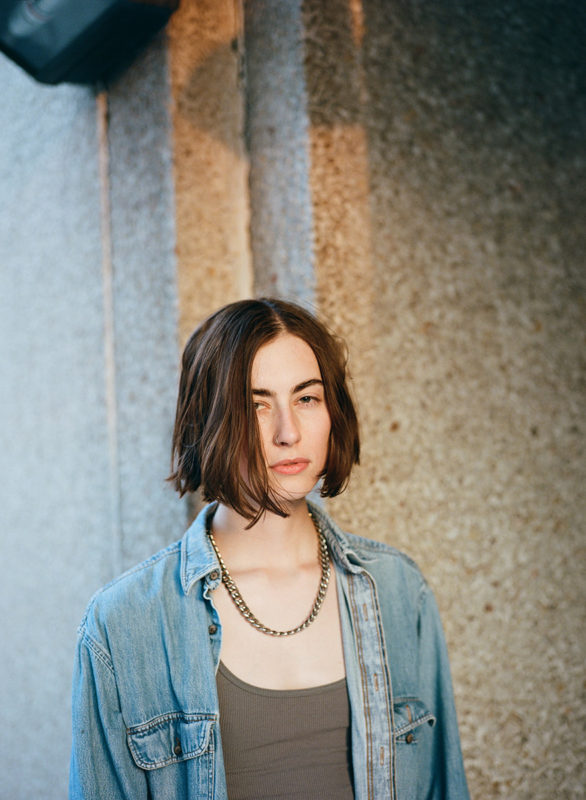 A woman with short brown hair wearing a light jean coat, green shirt, and necklace stands in front of a concrete wall.