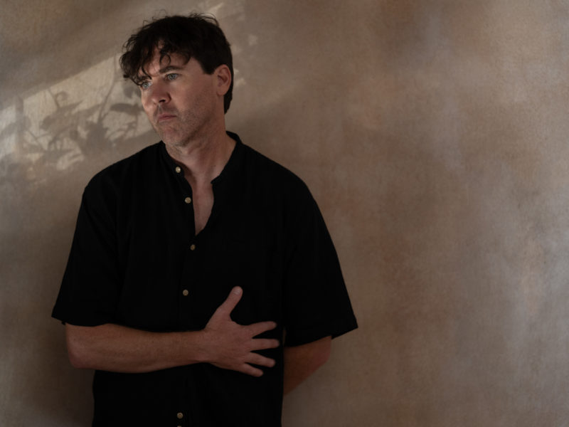 Color photo of Cass McCombs in a dark colored short sleeve shirt leaning up against a wall with his right arm crossing his mid-section.