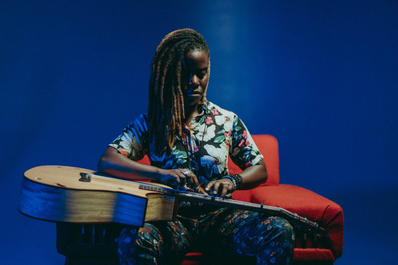 Yasmin Williams sits on a red chair in front of a blue background with her guitar on her lap. She is wearing a flowery shirt and pants.