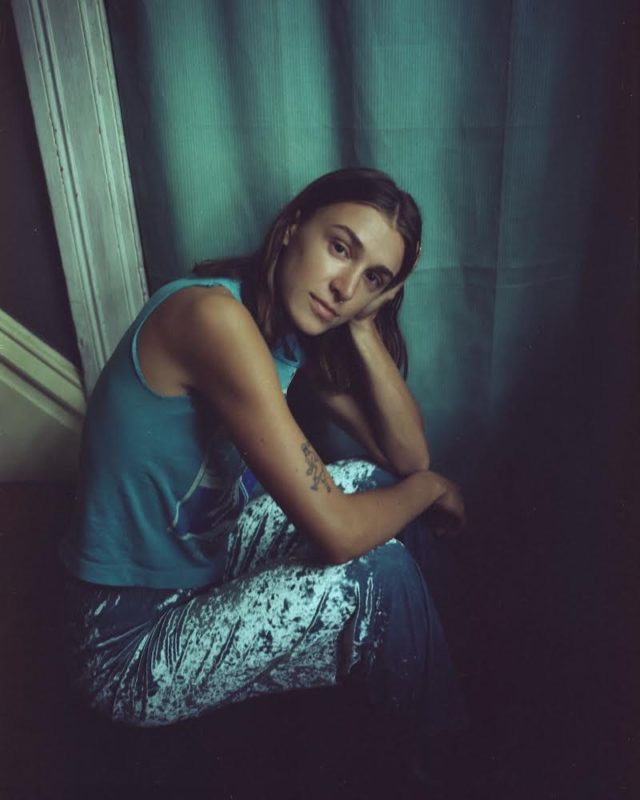 A person wearing a blue tank top is sitting with their arms on their knees and their head in their left hand.